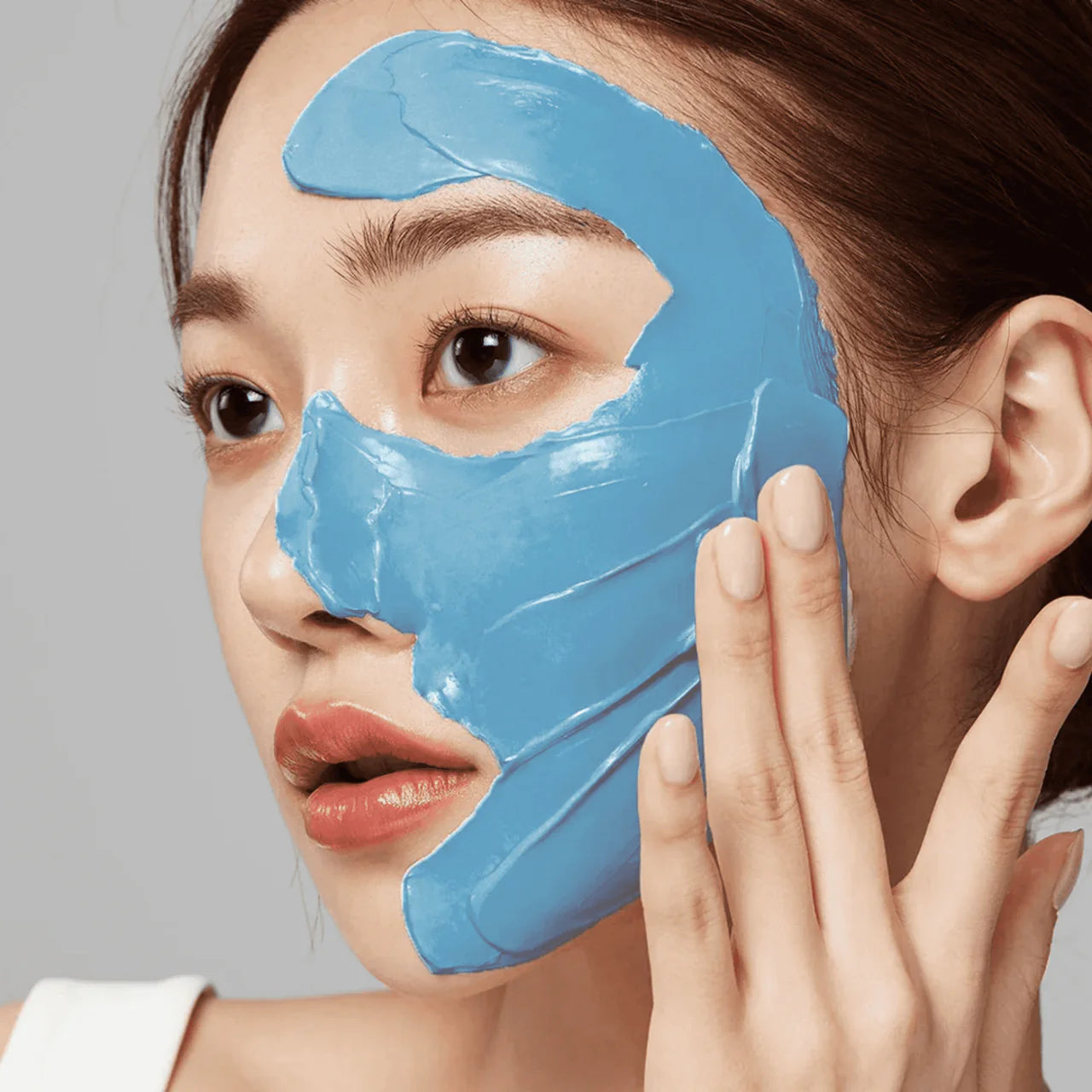 Woman applying a blue facial mask on her face against a gray background